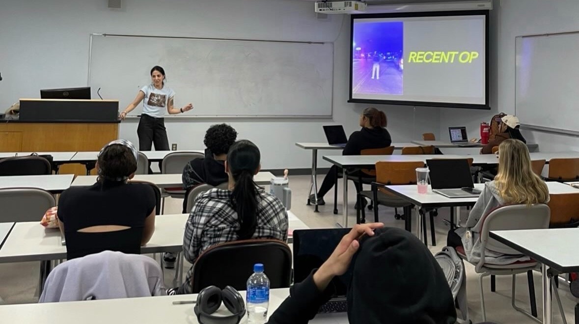 Roo Powell presenting to a university classroom, a slide reading Recent Op on the screen behind her